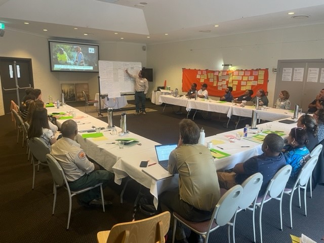 Participants seated at a ring of white desks facing a screen watch a presentation about the Aboriginal joint management model at the custodians workshop, in Ballina
