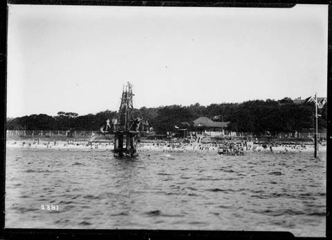 Black and white photo of the diving tower at Nielsen Park, Sydney Harbour National Park, featuring a tall structure overlooking the water, surrounded by trees and beachgoers enjoying the area.