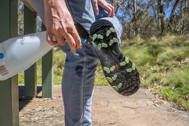 Someone spraying a clear liquid on the soles of their shoes. They are in the middle of a natural environment.