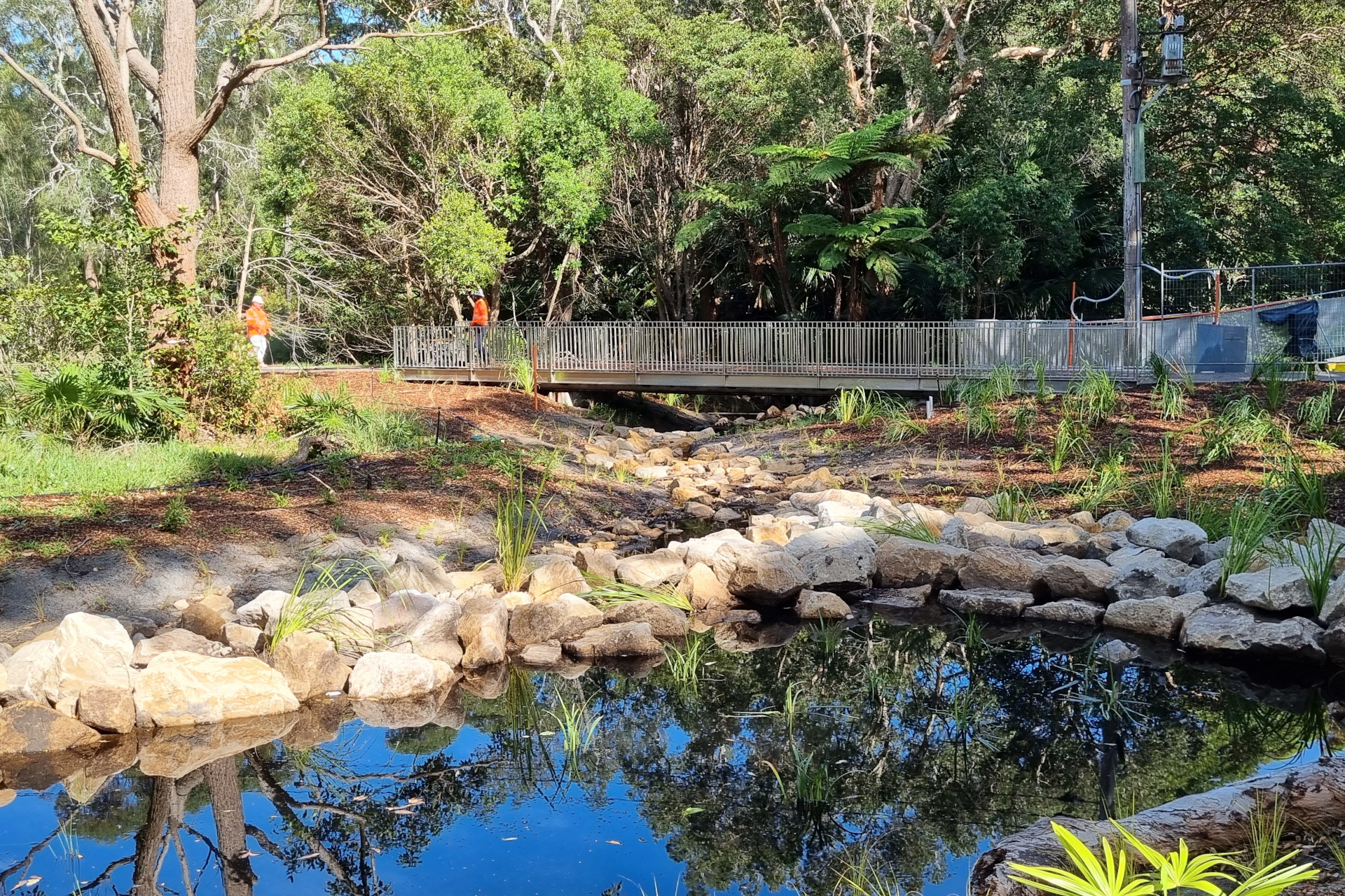 A small slow-flow pond area at Kurnell has clear blue water reflecting the surrounding trees and sky. The pond is bordered by rocks and lush greenery and there are trees behind it. In the background, there is a metal pedestrian walkway with railings going across Freshwater Stream. Two people wearing orange vests are in the background.