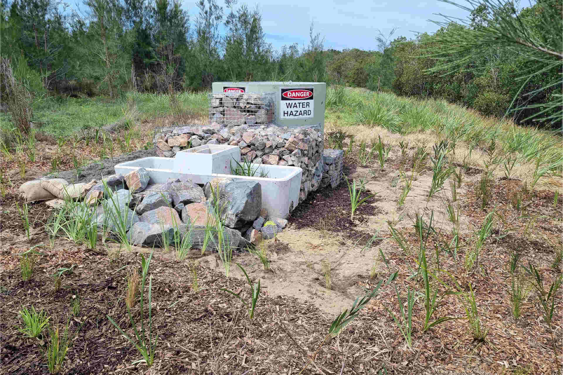 In a clearing is a tank and a tub surrounded by rocks and plants. The tank has a warning sign on it that says, 'Danger: Water hazard'