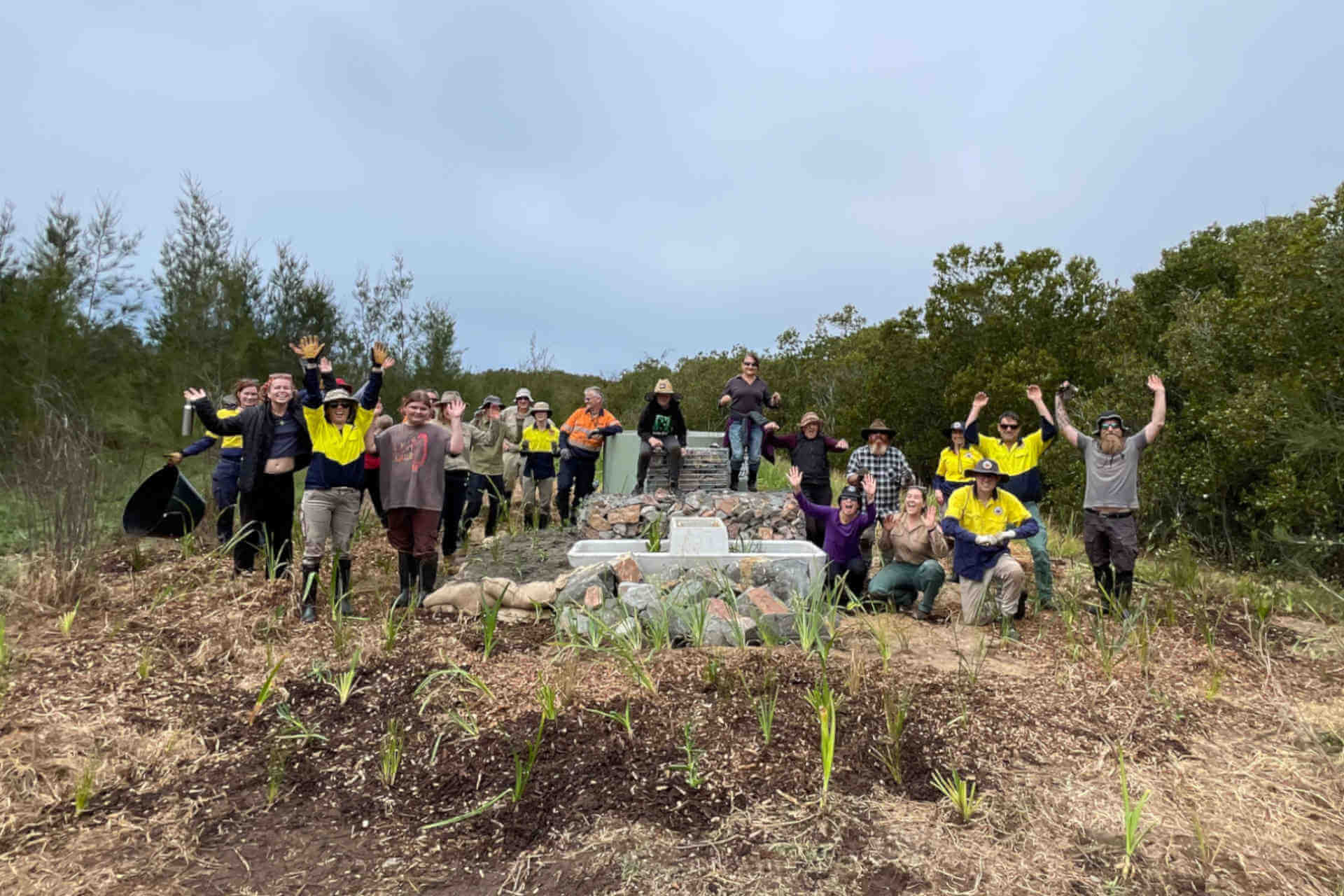 A group of around 20 people smile to the camera as they pose around an artificial pond and newly planted habitat in a clearing