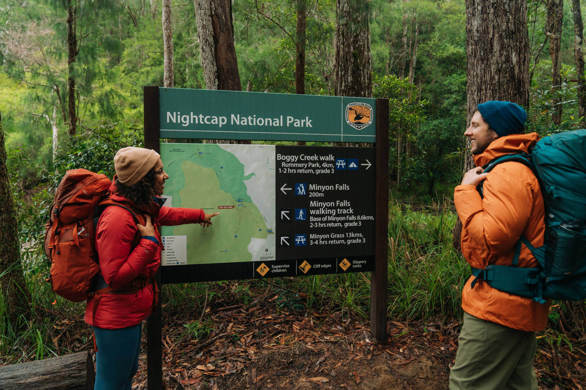 Two hikers, dressed in outdoor gear and carrying backpacks, stand in front of a signboard at Nightcap National Park. The sign provides information about two walking tracks: Boggy Creek walk (1-2 hours return, 4km) and Minyon Falls (4 hours return, 13km), both part of the Gidjuum Gulganyi Walk. One hiker points to a map on the sign. 