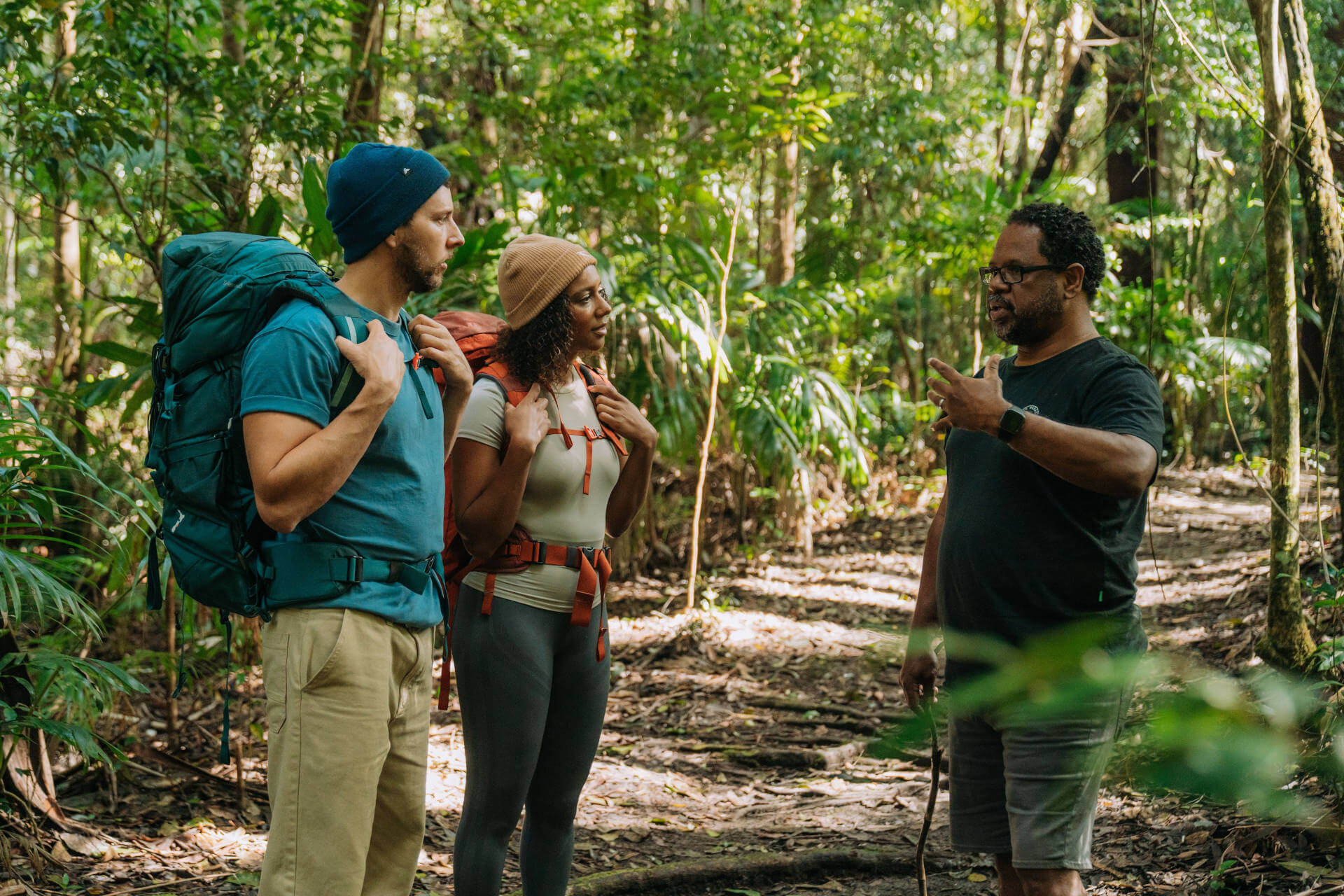 Three people are standing on a forest trail at Rummery Greek campground, Gidjuum Gulganyi Walk, engaged in conversation. Two of them are wearing backpacks and outdoor clothing, suggesting they are hiking. The third person appears to be giving directions or advice. 